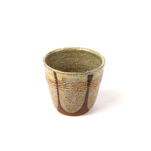 Puede incluir: Una taza de cerámica hecha a mano con una superficie texturizada y moteada. La taza presenta una base marrón claro con acentos verticales marrón oscuro y un interior verde claro. El borde tiene una banda amarilla clara. La taza mide unos 9 cm de alto.
