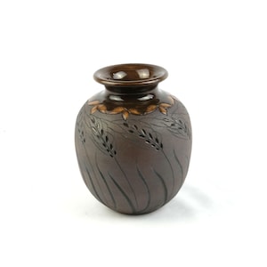 Könnte beinhalten: Eine dunkelbraune Keramikvase mit rundem Körper und ausgestelltem Rand. Die Oberfläche zeigt eingravierte Weizenhalmmuster im oberen Bereich, mit einer glatten, glänzenden Glasur am Hals und einer matten Textur am unteren Vasenkörper.