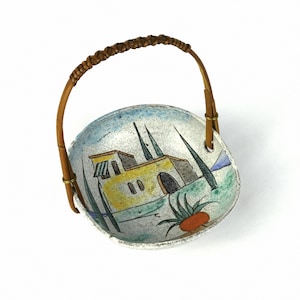 Puede incluir: Un cuenco de cerámica blanca con una escena pintada de una casa amarilla con árboles verdes y un cielo azul. El cuenco tiene un asa de bambú marrón.