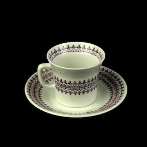 May include: A white teacup and saucer set with a decorative brown pattern. The cup has a handle and the saucer is round. The pattern is a repeating design around the rim of the cup and saucer.
