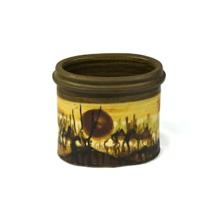 May include: A cylindrical ceramic container with a hand-painted desert scene. The design features a setting sun, camels, and silhouettes in shades of brown, yellow, and orange. The rim and base are a darker brown.