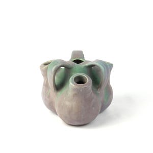 Puede incluir: Un jarrón de cerámica con un diseño único. El jarrón tiene una forma bulbosa con múltiples aberturas y asas. Presenta una mezcla de tonos verdes y grises-púrpuras apagados, creando una apariencia texturizada. La estética general es artística y decorativa.