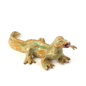 May include: A ceramic sculpture of a green and brown lizard with a textured surface, holding a small brown stick in its mouth.