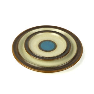 May include: A stack of three ceramic plates with concentric circles in brown, cream, and teal. The plates are arranged in a nested fashion, with the smallest plate on top. The plates have a simple, modern design.