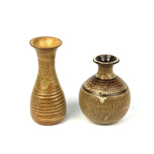 May include: Two ceramic vases in shades of brown and tan. The taller vase has a slender, cylindrical shape with horizontal ridges. The shorter vase is round with a narrow neck. Both have a rustic, handcrafted appearance.
