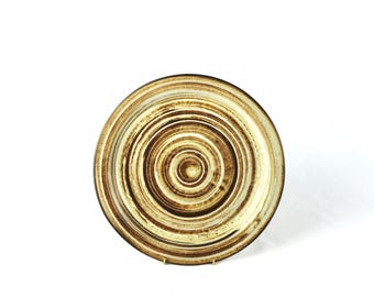 May include: A round ceramic plate with a concentric circle design in shades of brown and beige.