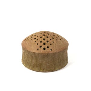 May include: A cylindrical brown ceramic flower frog with a domed top featuring a series of small, circular perforations arranged in a triangular pattern. The sides of the object have a vertical ribbed texture, and the base is flat and circular.