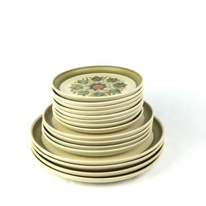 Può includere: Una pila di piatti in ceramica beige con bordo verde. Il piatto superiore presenta un motivo floreale con dettagli rossi, verdi e gialli. I piatti sono impilati, mostrando varie dimensioni. Lo sfondo è bianco.