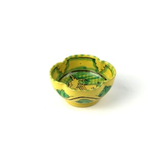 Op de afbeelding: Een kleine, gele keramische kom met een geschulpte rand. De kom heeft groen en zwart geschilderde accenten, waaronder een centraal beeld van een dier. De kom heeft een diameter van ongeveer 10 cm.