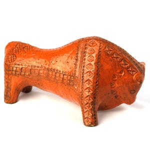 May include: Orange ceramic bull figurine with detailed carved patterns. The bull has a textured surface and is standing on four short legs. The figurine is a decorative item, likely for home decor.