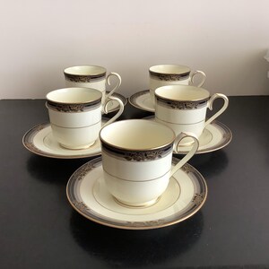 Op de afbeelding: Een set van vijf crèmekleurige kopjes en schotels. Elk kopje en schotel heeft een decoratieve band met donkergrijze en gouden bloemmotieven. De kopjes hebben gebogen handvatten en de schotels hebben een bijpassende rand.