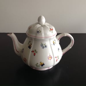 Könnte beinhalten: Weiße Keramik-Teekanne mit Deckel und Henkel. Die Teekanne ist mit kleinen, bunten Blumenmustern in Rot, Blau und Gelb verziert. Eine dünne rote Linie akzentuiert Ausguss und Henkel. Die Teekanne steht auf einer dunklen Oberfläche.