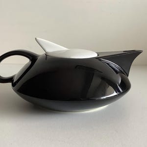 Könnte beinhalten: Eine schwarz-weiße Teekanne mit einem schlanken, modernen Design. Die Teekanne hat eine glänzende schwarze Oberfläche mit einem weißen Deckel und einem markanten Ausguss. Der Griff ist gebogen, und die Teekanne ist etwa 25 cm lang.