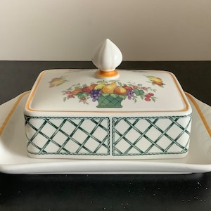 Pode incluir: Uma manteigueira de cerâmica branca com tampa com um desenho de cesta de frutas. A tampa tem um remate branco e é adornada com laranja. A base tem um padrão de treliça verde e uma borda laranja.