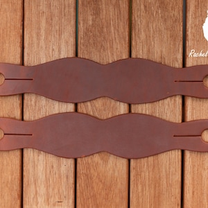May include: Two brown leather stirrup leathers with a single hole and a slit in each. The leathers are on a wooden surface. "Rachel Foil Custom Tack" is printed in white text on the right side of the image.