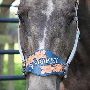 May include: Close-up of a horse wearing a custom leather halter. The halter is navy blue with the name "SMOKEY" in white script. Floral and leaf designs in tan and brown adorn the halter. The horse's face is gray with a white stripe.