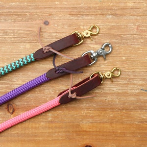 May include: Three leather dog leashes with different colored ropes. The leashes are brown leather with a brass hook on the end. The ropes are teal and brown, purple, and pink.