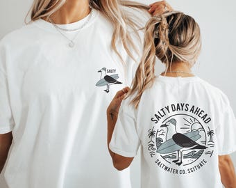 Könnte beinhalten: Weiße T-Shirts mit einem Möwen-Design. Ein Shirt hat eine kleine Möwe mit einem Surfbrett und dem Wort "SALTY". Das andere Shirt hat eine größere Grafik auf dem Rücken mit dem Text "SALTY DAYS AHEAD" und "SALT WATER CO. SCITUATE".