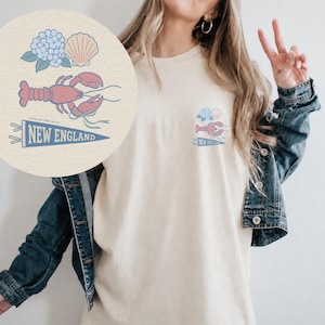 May include: Cream-colored t-shirt featuring a New England-themed graphic. The design includes a red lobster, blue hydrangea, and a seashell. The text "NEW ENGLAND" is displayed on a blue pennant. The shirt is worn by a person.