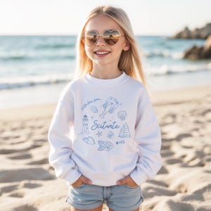 Op de afbeelding: Een witte sweatshirt met blauwe illustraties van een kreeft, schelpen, een vuurtoren en het woord "Scituate". De sweatshirt heeft ook de tekst "With love from" en het nummer "143". De persoon draagt hartvormige zonnebrillen.