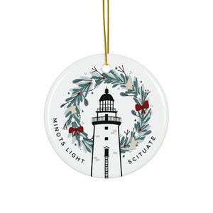 Op de afbeelding: Ronde witte keramische kerstversiering met een gouden ophangtouwtje. Het ontwerp toont een zwart-witte vuurtoren, omcirkeld door een groene krans met rode strikken. De woorden "Minots Light" en "Scituate" staan op de versiering.