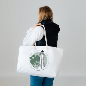 Op de afbeelding: Witte draagtas met een vuurtorenontwerp in groen en zwart, met de tekst "16 30" en andere tekst in een cirkel. De tas heeft witte touwhandvatten.