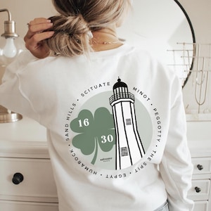 May include: White sweatshirt with a circular graphic featuring a lighthouse, a four-leaf clover, and the text "SCITUATE, SAND HILLS, MINOT, PEGGOTTY, THE SPIT, EGYPT." The clover has the numbers 16 and 30. The sweatshirt is a casual style.