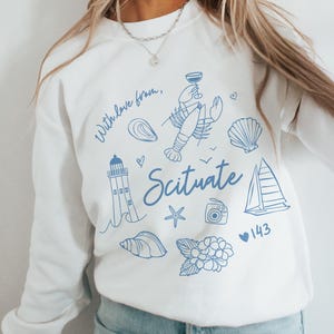 Op de afbeelding: Witte trui met een blauw grafisch ontwerp met een vuurtoren, kreeft, schelpen, een zeilboot, een camera, een ster en hortensia's. De tekst "With love from, Scituate 143" is ook in het ontwerp opgenomen.