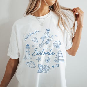 Op de afbeelding: Wit T-shirt met een blauw grafisch ontwerp met een vuurtoren, kreeft, schelpen, een zeilboot, een ster, een camera en bloemen. De tekst "With love from, Scituate 143" is op het shirt gedrukt.