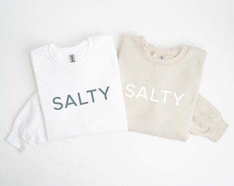 Könnte beinhalten: Zwei Sweatshirts, eines weiß und eines beige, werden gezeigt. Auf jedem Sweatshirt ist das Wort "SALTY" aufgedruckt. Der Text auf dem weißen Sweatshirt ist türkis, während der Text auf dem beigen Sweatshirt weiß ist. Die Sweatshirts sind gefaltet.