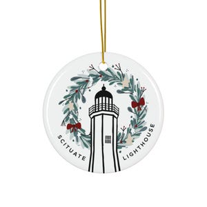Op de afbeelding: Witte, ronde keramische ornament met een zwart-wit vuurtorenontwerp. De vuurtoren is omcirkeld door een krans van groene bladeren en rode strikken. De tekst "SCITUATE LIGHTHOUSE" is rond de rand van het ornament gedrukt.