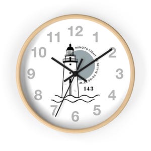 May include: A round wall clock with a light wood frame. The white face features a black and white lighthouse illustration, gray numerals, and black clock hands. The text "MINOTS LIGHT" and "143" are also visible.