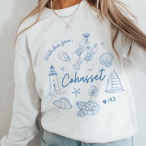 Op de afbeelding: Witte sweatshirt met blauwe illustraties van een vuurtoren, een kreeft en een zeilboot. Het woord "Cohasset" is cursief geschreven, met de zin "With love from," boven de illustraties.