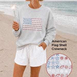 May include: Light gray crewneck sweatshirt featuring an American flag design made of blue stars and red and white shells. The text "American Flag Shell Crewneck" is visible. The person is wearing white shorts and holding a sparkler.
