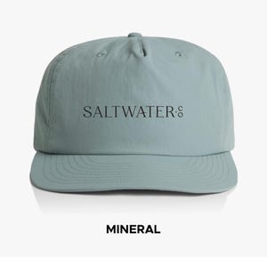 May include: A pale blue baseball cap with the word "SALTWATER." printed in black across the front. The word "MINERAL" is printed in bold black letters below the cap. The cap has a curved peak and a rounded crown.