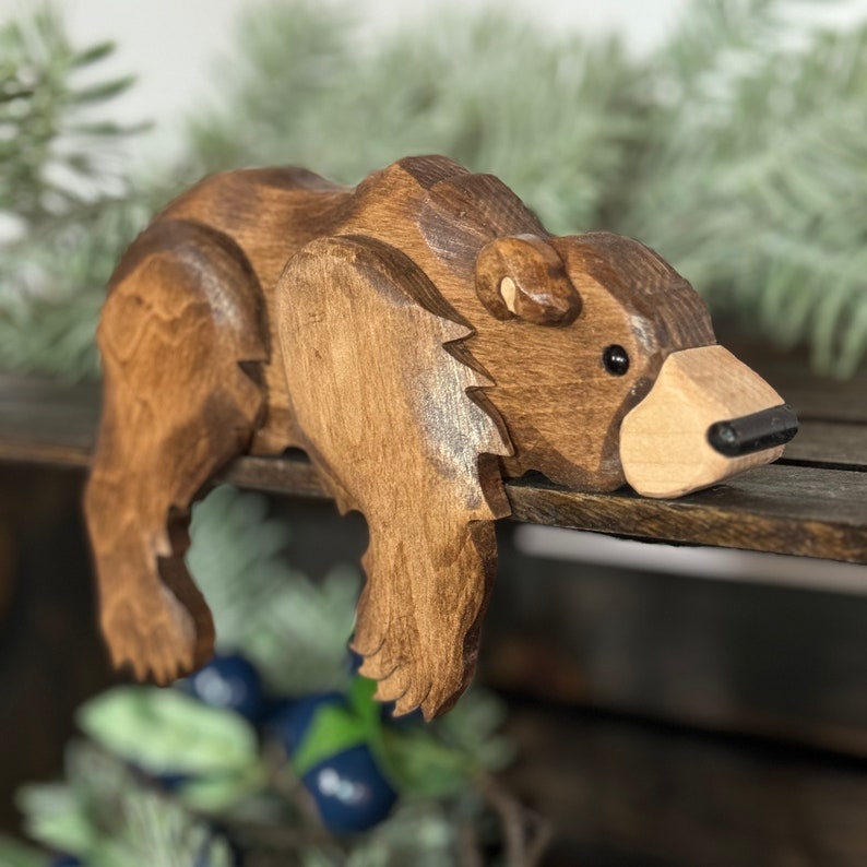 May include: A handcrafted wooden bear figurine in shades of brown. The bear is carved with detailed features, including a light-colored snout and black eyes. The bear is posed as if it is resting on a shelf. A charming decorative item.