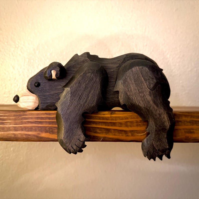 May include: A hand-carved wooden bear sculpture in a relaxed pose, painted black with a light-colored snout and ear. The bear is resting on a stained wooden shelf, showcasing the natural wood grain. A rustic, decorative item.