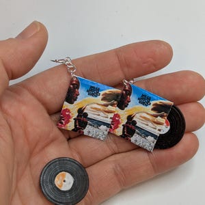 Puede incluir: Un par de pendientes con discos de vinilo en miniatura con la portada del álbum "Miles Davis - Bitches Brew". Los pendientes son plateados y tienen un cierre de gancho. También hay un solo disco de vinilo en miniatura sobre una superficie blanca.