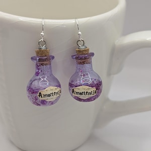 May include: A pair of earrings shaped like small, purple potion bottles with cork stoppers. The bottles are filled with purple glitter and have a label that reads "Amortentia". The earrings hang from silver-colored hooks.
