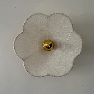 Op de afbeelding: Een bloemvormige wandlamp met een textuur, off-white stoffen kap. Het midden heeft een goudkleurig, bolvormig accent. De lamp is ontworpen om aan de muur te worden bevestigd en zorgt voor sfeerverlichting.