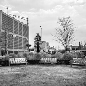 May include: Black and white photograph of a public park with benches arranged in a circle. A large clock tower stands in the background, along with a multi-story building and a cloudy sky.