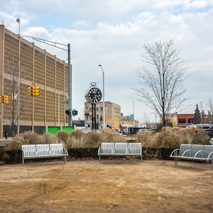 May include: An outdoor scene featuring several silver metal benches arranged in a circle. A large clock tower stands in the background, along with a tall, brown building and a cloudy sky. The ground is covered in brown dirt and dry grass.