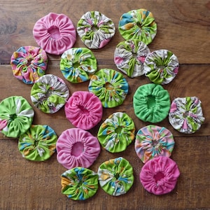 May include: A selection of handmade fabric yo-yos in various colours and patterns. The yo-yos are mainly pink and green, with floral and abstract designs. They are arranged on a wooden surface.