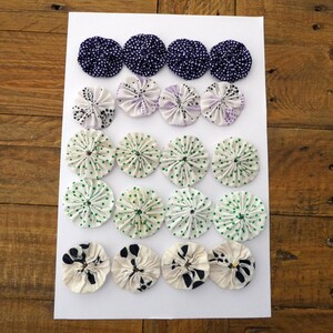 May include: A collection of fabric yo-yos in different colours and patterns displayed on a white surface. The yo-yos feature designs in navy blue, lavender, green, and black. The yo-yos are made of fabric and are circular in shape.