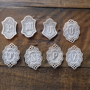 May include: Eight white lace appliques with embroidered letters, including P, M, U, T, N, P, and V. The appliques are in various shapes, including shields and ovals.
