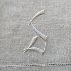 May include: A light beige linen fabric featuring a white embroidered letter S in a gothic script style. The embroidery has a raised, textured appearance against the woven surface of the textile, which includes a visible hemmed edge at the bottom.