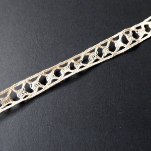 May include: Close-up of a cream-colored lace trim with an openwork design. The trim features a repeating pattern of interconnected shapes, creating a delicate and airy texture. The lace is set against a dark background, highlighting its intricate details.
