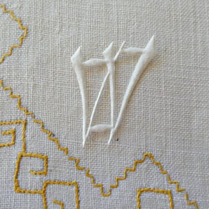May include: A piece of off-white fabric featuring white raised embroidery in the shape of the letter W, accompanied by a geometric pattern stitched in bright yellow thread that forms a series of connected angular lines across the textile surface.