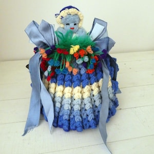 May include: A handmade, whimsical doll with a blue and white crocheted body, adorned with colorful yarn flowers and a blue ribbon. The doll has a small, light blue face and a matching hat. The doll is approximately 15 cm tall.
