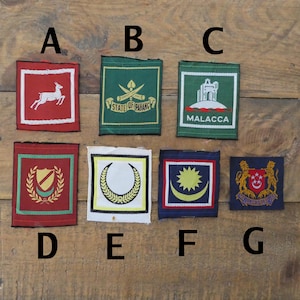 May include: Seven embroidered patches with diverse designs and colours. The patches display images like a deer, a shield, and a crescent moon. Some patches include text, such as "MALACCA" and "STATE OF PAHANG".